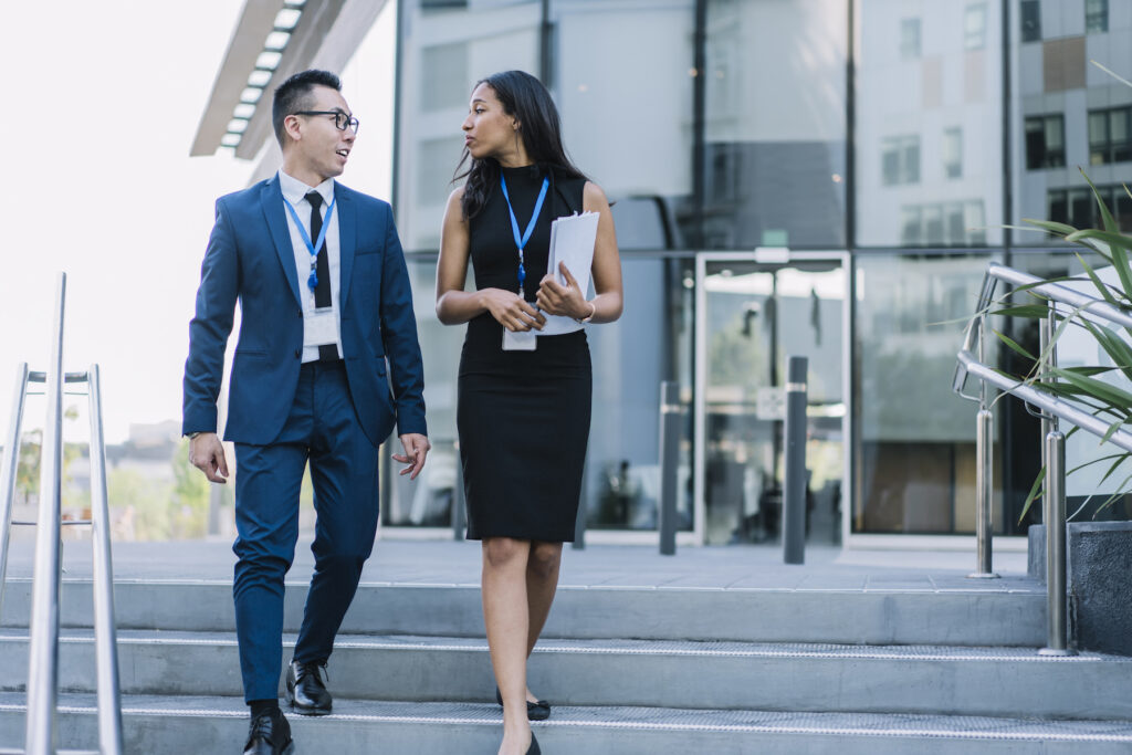 Young elegant African American businesswoman with Asian male colleague walking on street stairway carrying papers while meeting on modern conference