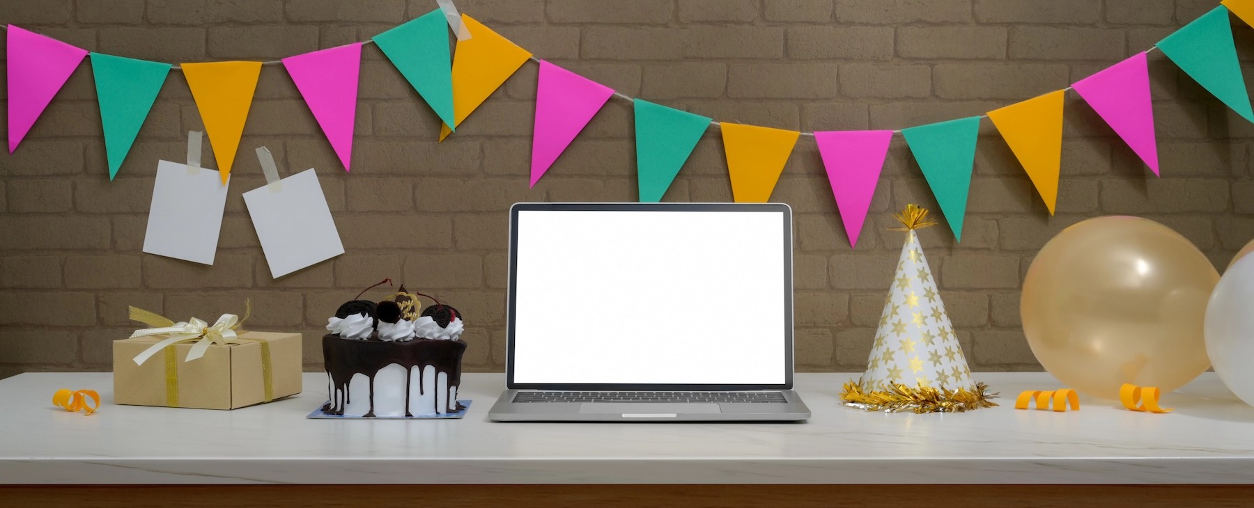 Online birthday party concept with mock-up laptop, Black Forest cake, present box and decorations on white desk and brick wall.