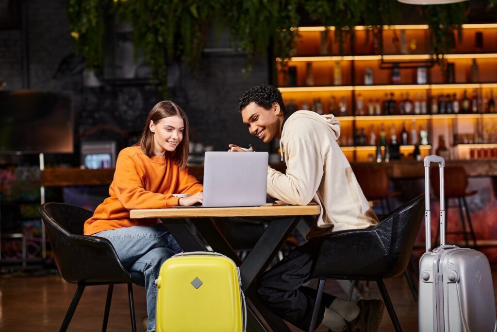 Black man and woman looking at laptop on cafe table, with their luggage beside them.
