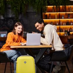 Black man and woman looking at laptop on cafe table, with their luggage beside them.