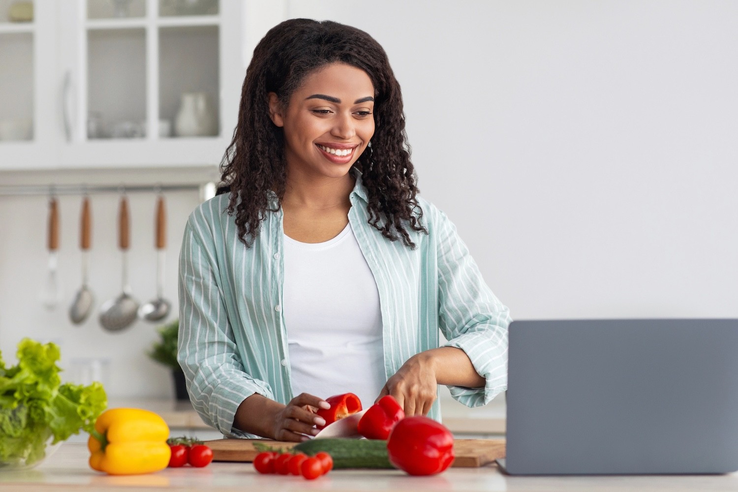  Smiling millennial African American female cuts pepper and looks at laptop in kitchen interior with bright vegetables on table.