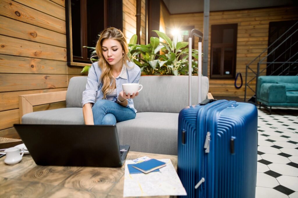 Young businesswoman on computer, holding a cup of coffee, with a suitcase beside her.