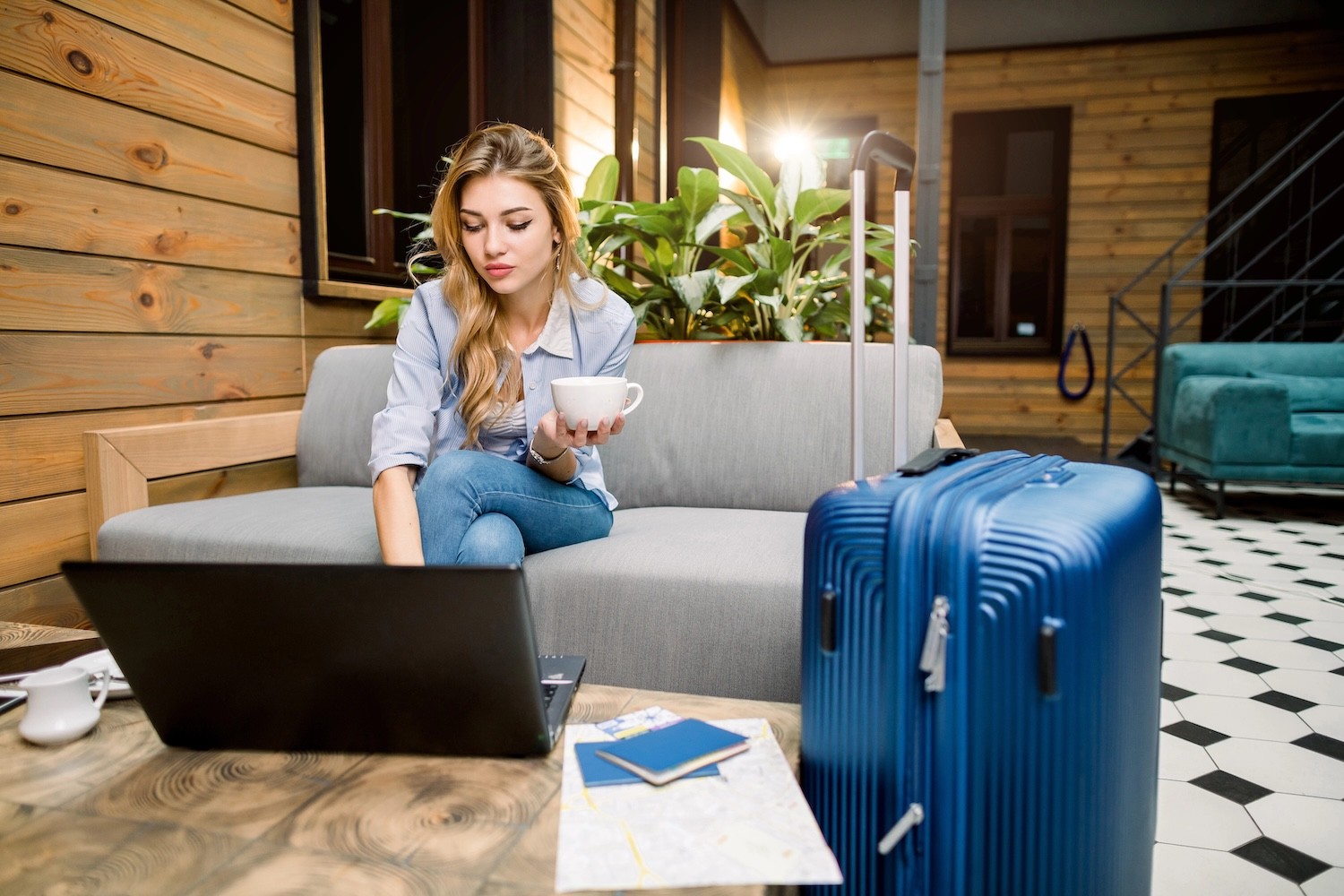 Young businesswoman on computer, holding a cup of coffee, with a suitcase beside her. 