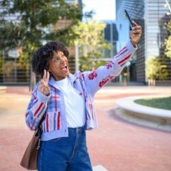 Joyful young woman making peace sign selfie in city park.