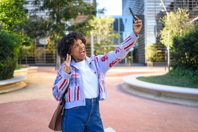 Joyful young woman making peace sign selfie in city park.