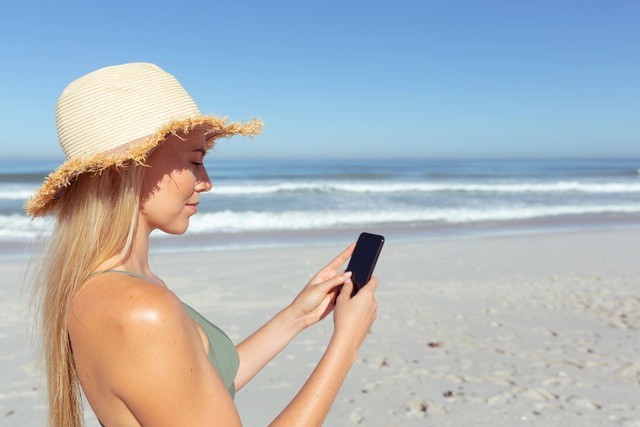 Woman on the beach on a sunny day, wearing a hat, and looking at her smartphone. 
