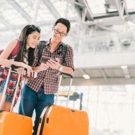 Asian couple travelers using smartphone checking flight or online check-in at airport, with passport and luggage. Air travel or mobile phone technology concept