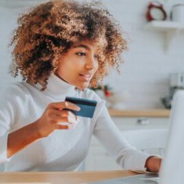 Lady with perfect dark skin holding credit card and inserting bank information for online shopping sits near laptop closeup