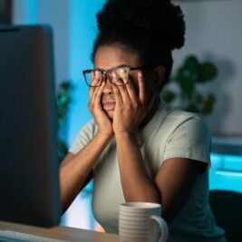 Symptoms of overworking. Young tired overworked african woman freelancer in eyeglasses working late from home, sitting in front of monitor, feeling eye strain and fatigue during computer work.