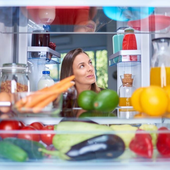 Confused hungry woman standing in front of opened fridge full of groceries.