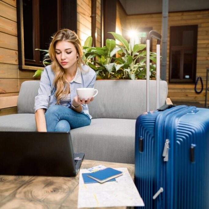 Young businesswoman on computer, holding a cup of coffee, with a suitcase beside her.