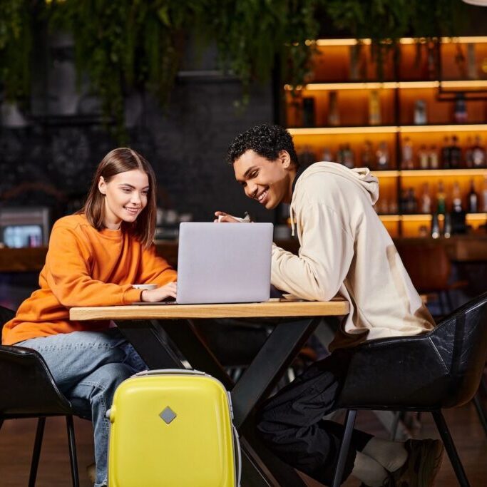 Black man and woman looking at laptop on cafe table, with their luggage beside them.