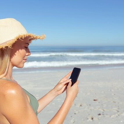 Woman on the beach on a sunny day, wearing a hat, and looking at her smartphone.